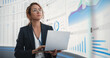 © Gorodenkoff - In the Monitoring Office: Successful Hispanic Businesswoman Holding Laptop Computer And Analyzing Data On Big Digital Screen With Graphs and Charts. Female IT Entrepreneur Running Software Company.