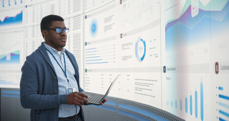 Fototapete - Confident Black Businessman Holding Laptop And Reviewing Financial Reports On Big Digital Screen In Monitoring Office. Male Entrepreneur Analyzing Growth And Profit Margins Of Tech Start Up Company.