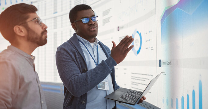 African Data Science Team Lead With Laptop Computer Reporting To Indian CEO About Growth And Sales In Front Of Big Digital Wall In The Office. Two Successful Men Discussing Performance Of The Company.