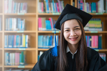 Wall Mural - Portrait young happy and excited Asian woman university graduate in graduation gown and cap in the library. Education stock photo