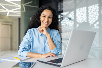 Portrait of young beautiful business woman inside office at workplace with laptop, smiling successful hispanic woman smiling and looking at camera, satisfied female boss in shirt sitting at table.