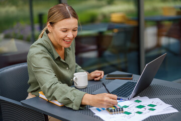 Wall Mural - Smiling businesswoman working on laptop and making notes in cafe terrace while drinking coffee