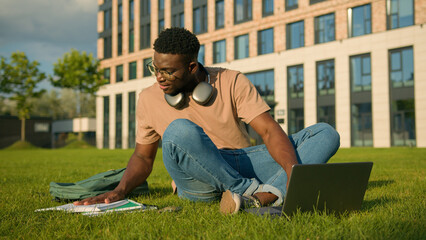 Happy smiling African American man university college academy student guy teenager male studying homework online on grass meadow in park study learning with book copybook laptop prepare for exam study