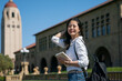 attractive asian Chinese female exchange student holding book and pushing hair back while looking at camera with a smiling face at school university with hoover tower at background