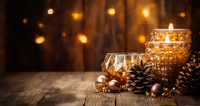 Festive Holiday Scene With Candles And Decorations. Two Lit Candles In Glass Holders With Gold Beads. Pine Cones And Baubles On A Wooden Table. Wooden Wall With Fairy Lights In The Background. 