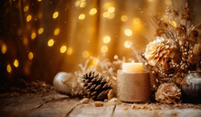 Candle In A Gold Glittery Holder On A Wooden Surface With Dried Flowers And Pine Cones. The Background Is A Wooden Wall With String Lights. The Lights Are Yellow And Give The Image A Warm Glow.