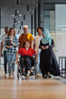 © .shock - A diverse group of young business people walking a corridor in the glass-enclosed office of a modern startup, including a person in a wheelchair and a woman wearing a hijab, showing a dynamic mix of