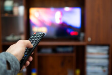 Hand Of A Person In The Foreground Holding A Remote Control And Pointing To A TV In The Background Out Of Focus.