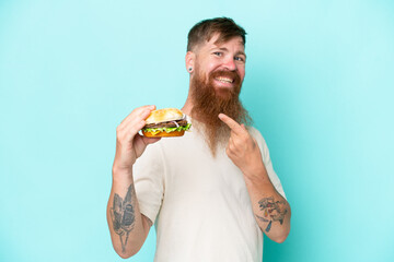 Wall Mural - Redhead man with long beard holding a burger isolated on blue background and pointing it