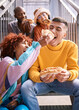 © Wesley/peopleimages.com - Friends, city and feeding food outdoor for happiness, funny laugh and fun on stairs. Diversity, eating and gen z group of men and women with a hotdog on a date, adventure and freedom in urban town