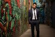 A suave, sharply-dressed businessman stands confidently in front of a vibrant, graffiti-covered wall, exuding an air of rebellious sophistication.
