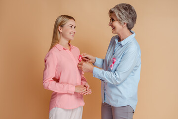 Wall Mural - Smiling women attach pink ribbon looking each other isolated on beige background. Health care, support, solidarity, prevention. Breast cancer awareness month concept