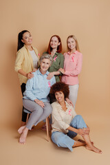 Wall Mural - Group of smiling multiethnic women wearing breast cancer pink ribbon support each other celebrate recovery isolated on beige background. Health care, prevention. Breast cancer awareness month