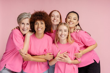 Group of smiling confident multiracial women wearing t shirts with pink ribbon looking at camera isolated on pink background. Health care, support, prevention. Breast cancer awareness month concept