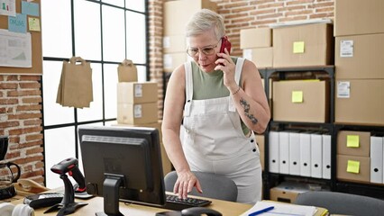 Wall Mural - Middle age grey-haired woman ecommerce business worker talking on smartphone using computer working at office