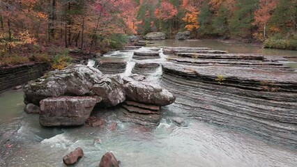 Wall Mural - Multiple waterfalls flow over rock cascade in the mountains during autumn with fall colored forest tree leaves via panning aerial  