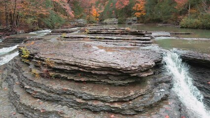Wall Mural - Multiple waterfalls flow over rock cascade in the mountains during autumn with fall colored forest tree leaves reverse drone flight   