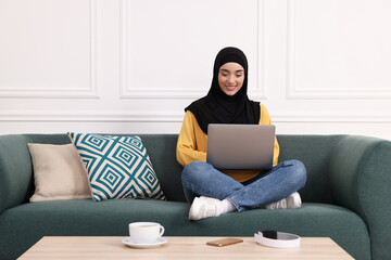 Poster - Muslim woman in hijab using laptop on sofa indoors. Space for text