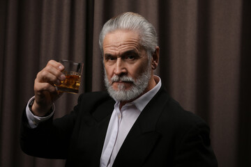 Canvas Print - Senior man in formal suit holding glass of whiskey with ice cubes on brown background