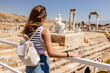 A young woman in a striped top and blue denim shorts with an off white backpack looks at a picturesque landscape of the ruins of an ancient city. Hierapolis, Pamukkale, Türkiye - July 29, 2023