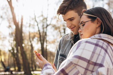 Young loving couple standing in the city park in sunny weather looking at phone screen, hugging smiling kissing laughing spending time together. Autumn, fall season, orange yellow red maple leaves

