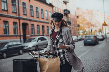 Wall Mural - Young caucasian woman using a smart phone while pushing her bicycle on a city sidewalk