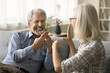 © fizkes - Positive senior husband with hearing disorder, disability talking with hands to mature wife, using finger signs for conversation, speaking gesture language, smiling, laughing, sitting on sofa