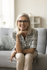 Wall Mural - Cheerful beautiful senior lady in trendy glasses leaning chin on hand, sitting on home sofa, looking at camera with toothy smile, talking on online video conference call. Vertical shot