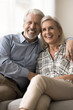 © fizkes - Cheerful positive senior retired husband and wife hugging on home sofa, looking at camera with toothy smiles, showing healthy white teeth, promoting elderly dental service. Vertical portrait shot