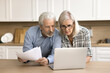 © fizkes - Serious senior couple checking domestic paper documents at kitchen table, using online financial banking application on laptop, doing household accounting paperwork