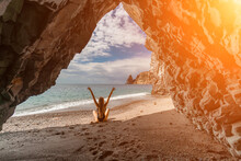 Woman Travel Sea. View Of A Woman In A Black Swimsuit From A Sea Cave Attractive Woman Enjoying The Sea Air Sits On The Beach And Looks At The Sea. Behind Her Are Rocks And The Sea