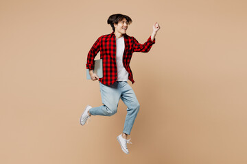 Full body young IT man of Asian ethnicity he wear red shirt casual clothes jump high hold closed laptop pc computer hurry up run fast isolated on plain pastel light beige background Lifestyle concept