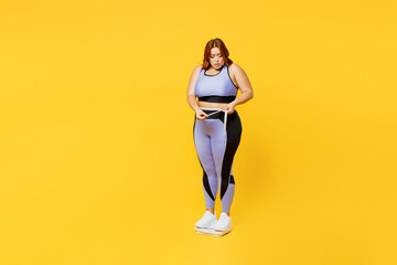 Wall Mural - Full body sad young plus size big fat fit woman wear blue top warm up train stand on scales check result look at measure tape isolated on plain yellow background studio home gym Workout sport concept.