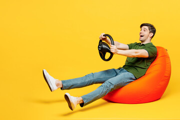 Full body side view excited young happy man wears green t-shirt casual clothes sit in bag chair hold steering wheel driving car isolated on plain yellow background studio portrait. Lifestyle concept.