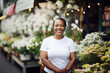 A smiling middle-aged afro woman in a white t-shirt is standing on the blurred background of a flower shop. The concept of her own business. Mock-up for design. Blank template. Al generated