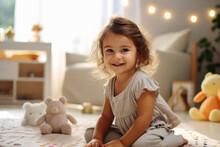 Full-body Shot Of A Happy 3-year-old Girl Playing On The Carpet In The Living Room With Toys Around. She Is Wearing A Light Beige Home Outfit