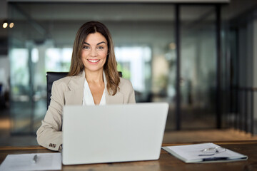 Happy smiling mature middle aged professional business woman manager executive or lawyer looking at camera at workplace, working on laptop computer technology in office sitting at desk, portrait.