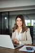 © insta_photos - Happy mature mid aged professional business woman manager executive, lawyer attorney looking at camera at workplace working on laptop computer technology in office sitting at desk, vertical portrait.