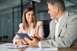 © insta_photos - Two professional mature executive managers business team Latin man and woman wearing suits talking working in office at meeting having conversation using tablet discussing corporate digital solutions.