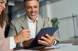 © insta_photos - Smiling older professional executive business man talking working in corporate office at meeting with female bank manager consulting client looking at digital tablet computer discussing sales project.