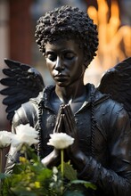 Sculpture Of Black Male Wings Angel Surrounded By Flowers And Garden Plants