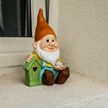 A Gnome Sits On The Windowsill Beside A Green Birdhouse