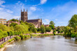 Worcester Cathedral and the River Severn on a bright sunny summer day. Swans on the river, people strolling on riverside path, leafy green trees, and cathedral towering over all. 