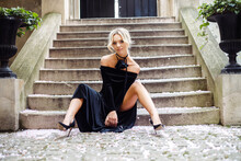 Portrait Of Young Attractive Woman Wearing Black Long Velvet Dress, High-heeled Shoes Sitting On Old Concrete Stairs.