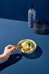 Wall Mural - Asian soup with mushrooms, seafood, and egg, with a woman's hand holding and dipping a spoon on a solid blue background