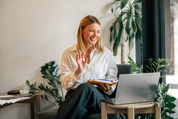 Wall Mural - Smiling business woman talking with client via videocall and waving hi while sitting in coworking