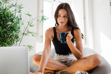 Smiling Young Woman With Coffee Cup Using Laptop In Apartment