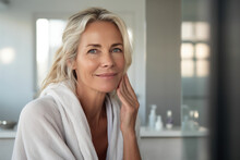 Headshot Of Gorgeous Mid Age Adult 50 Years Old Blonde Woman Standing In Bathroom After Shower Touching Face, Looking At Reflection In Mirror Doing Morning Beauty Routine. Older Skin Care Concept.