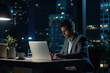 © AI_images - Young busy Latin business man executive working on laptop at night in dark corporate office. Professional businessman manager using computer sitting at table, big city evening view