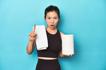 Young Asian sportswoman with protein bottle, gym setup, having an idea, inspiration concept.